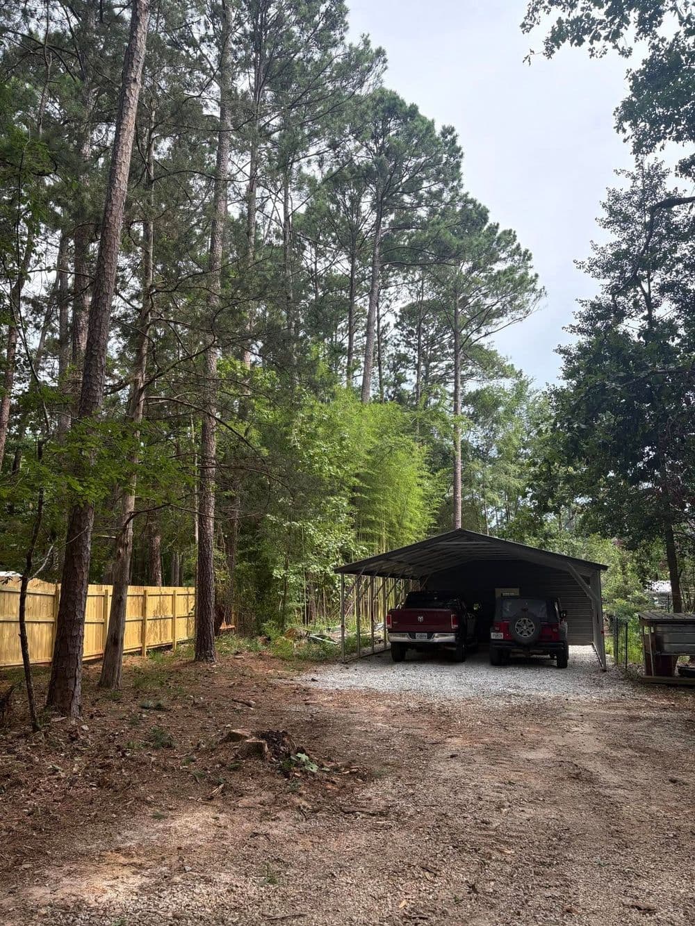 Two vehicles parked under a carport in a wooded area with tall pine trees.