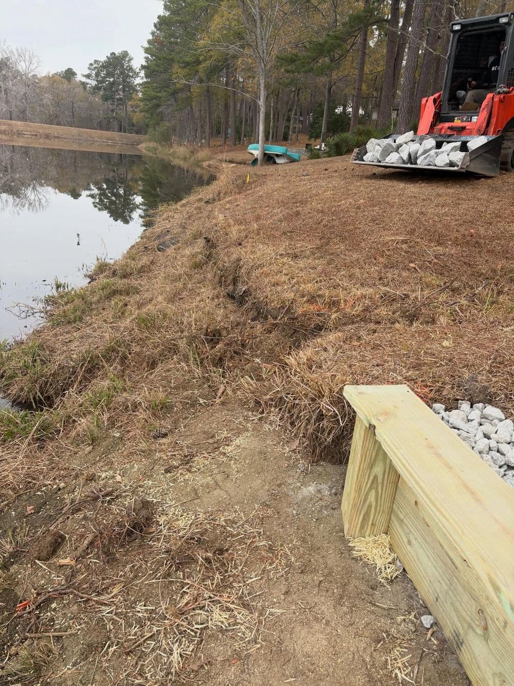 Lakeside landscaping with a wooden bench, gravel, and construction equipment near a pond.