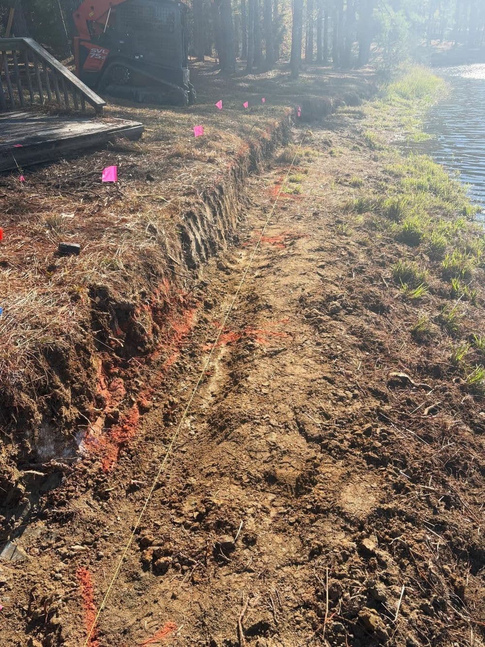 Construction site near a pond, marked by flags and excavation along a dirt path.