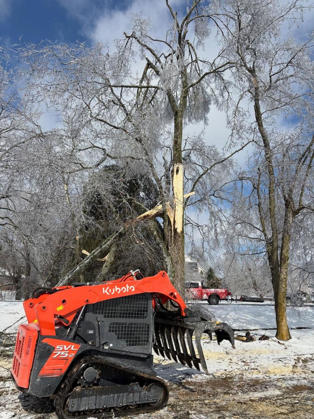 Kubota skid steer loader with grapple attachment near ice-covered trees after winter storm.